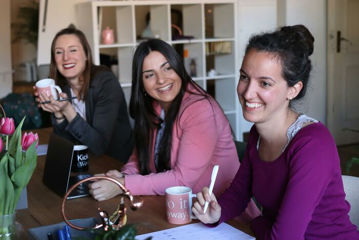 Three women smiling and chatting around a table, illustrating moments from adult life feeling like being picked last in gym class.