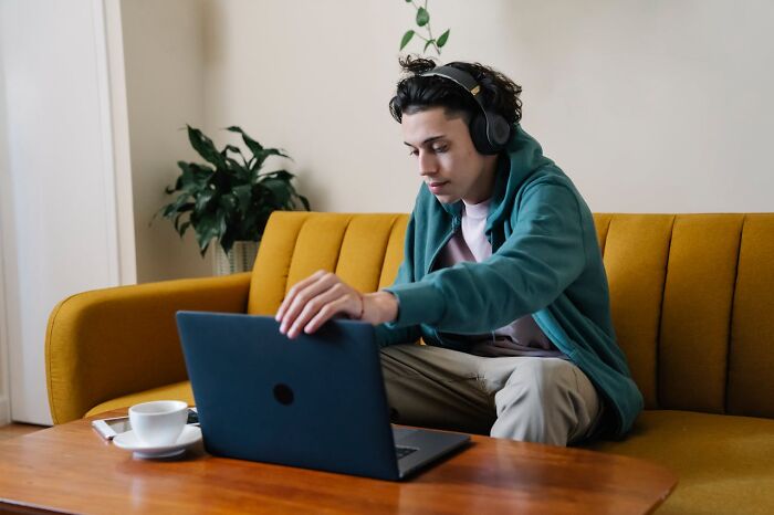 Young man wearing headphones sitting on a couch, using laptop, illustrating excessively lazy thing shared by people.