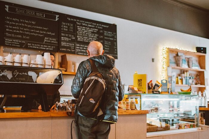Customer wearing a backpack orders coffee at a café counter illustrating real-life cheat codes people swear by for success.