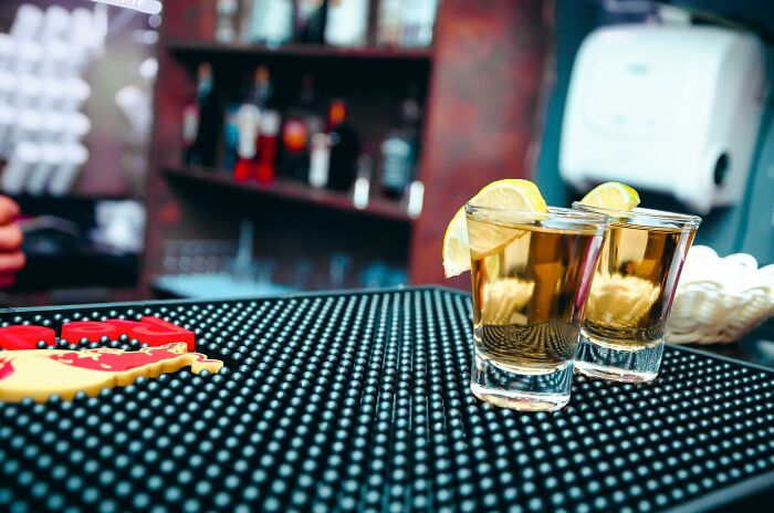 Two shot glasses with lemon wedges on a bar mat inside a dimly lit bar behind the tap with bartenders.