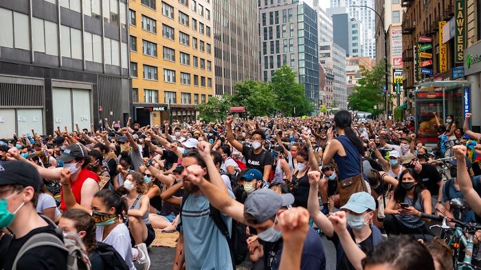 Large diverse crowd wearing masks raising fists during a middle class protest in an urban city street setting.