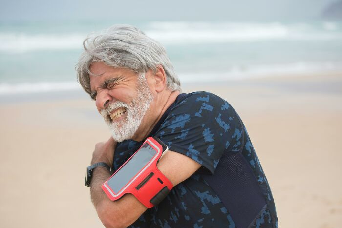 Older man in workout gear grimacing in pain holding his shoulder on a beach illustrating harsh life advice lemons