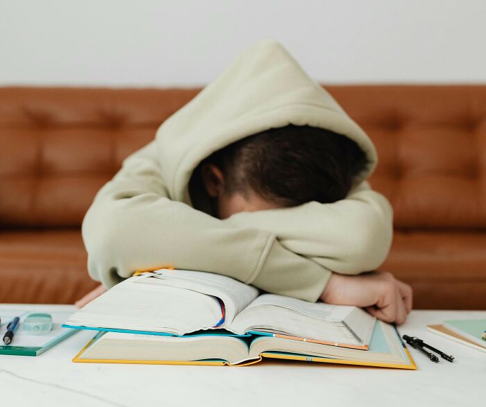 Teen in hoodie resting head on arms at table with open books, reflecting on soul-crushing things parents told them.