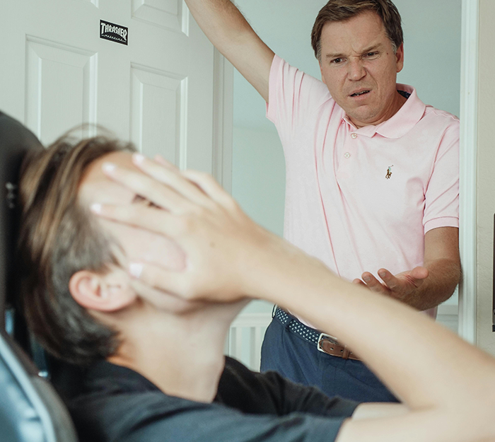 Man in pink shirt angrily confronts teen covering his face in a room, depicting tension over mom&rsquo;s new boyfriend issues.