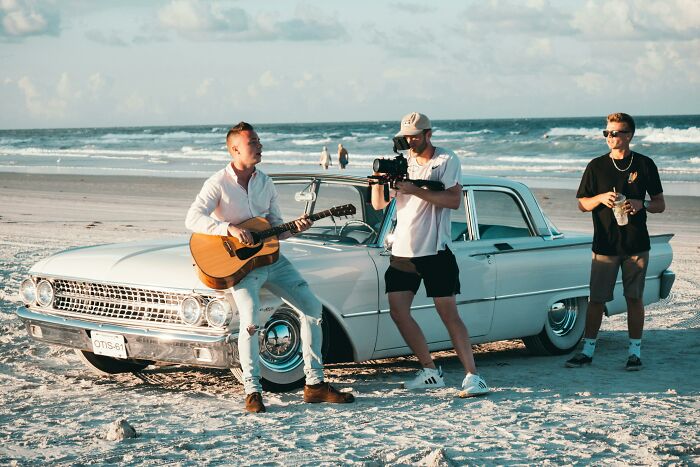 Musician playing guitar beside vintage car on the beach while a videographer films an iconic music video scene.