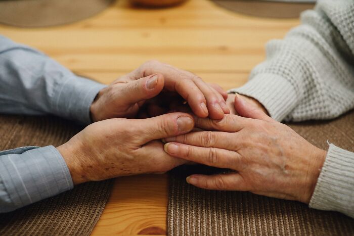 Two pairs of older hands gently holding each other on a wooden table showing body changes that indicate aging.