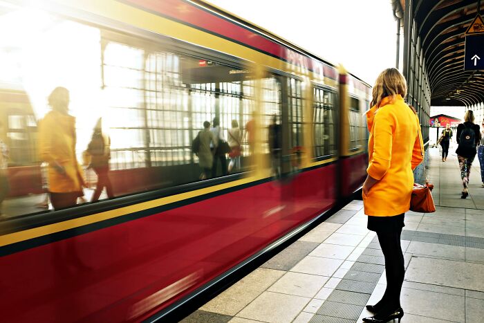 Woman in bright orange coat standing on platform as red train arrives, illustrating cultural differences in urban transportation.