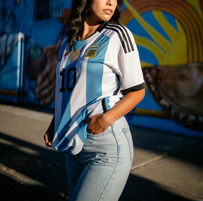 Woman wearing Argentina soccer jersey standing outdoors near a colorful mural, representing women asking questions to men.
