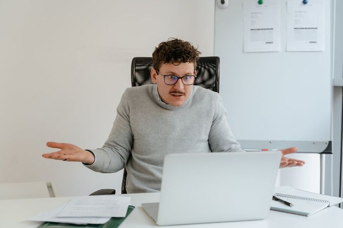Man in glasses and gray sweater looking confused while working on laptop, illustrating weird finds shared by doctors and morticians.