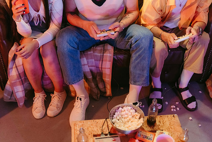 Three people sitting closely on a couch playing video games with snacks and drinks, family in need scene.