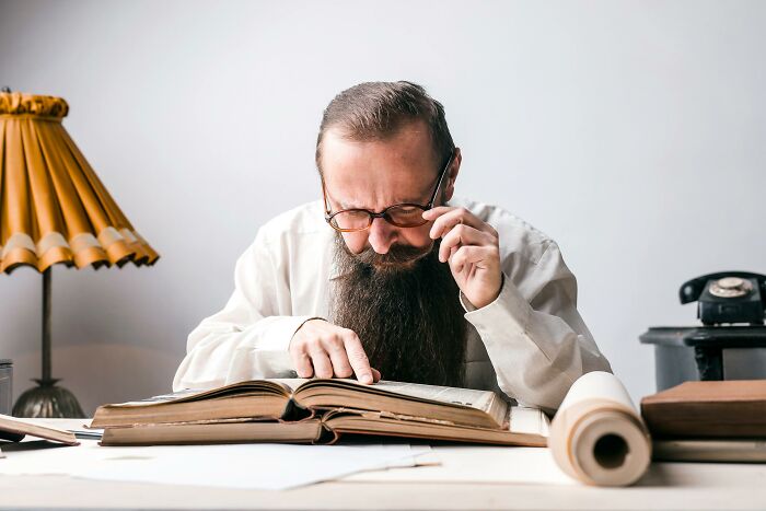 Man with glasses reading a large book intently, studying world history key events at a desk with vintage lamp and phone. Man with glasses reading a large book intently, studying world history key events at a desk with vintage lamp and phone.
