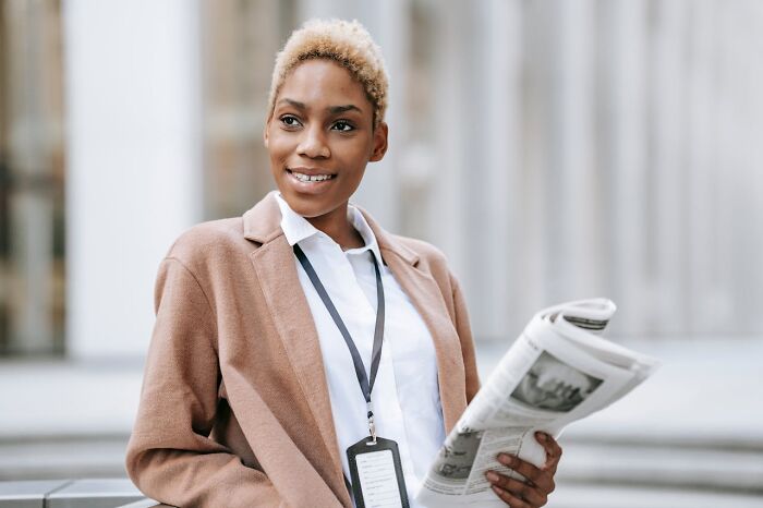 Young professional woman smiling and holding a newspaper outdoors, illustrating real-life cheat codes that work almost every time.