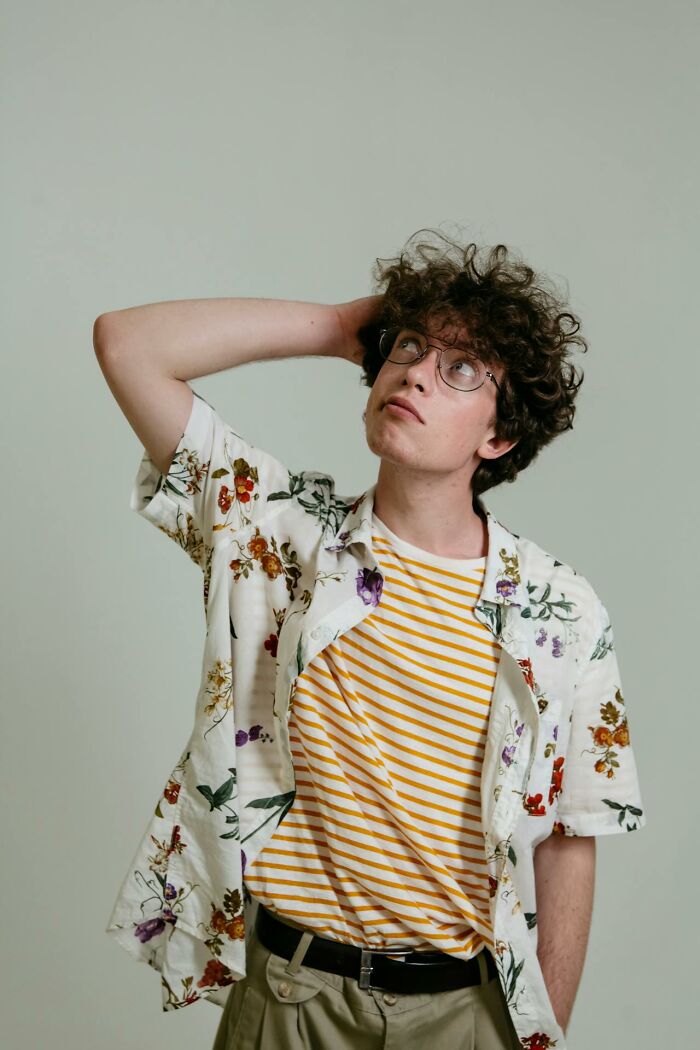 Young Gen Z man with curly hair and glasses, wearing a floral shirt and striped tee, reflecting on middle class challenges.