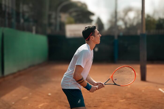Young man on a clay tennis court holding a racket, focused and ready to play in an outdoor sports setting.