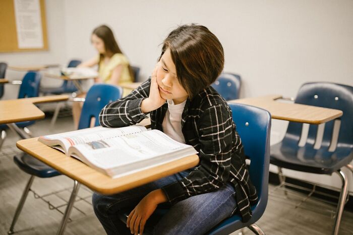 Student in classroom reading textbook focused on learning with unhinged but effective teacher strategies visible in background.