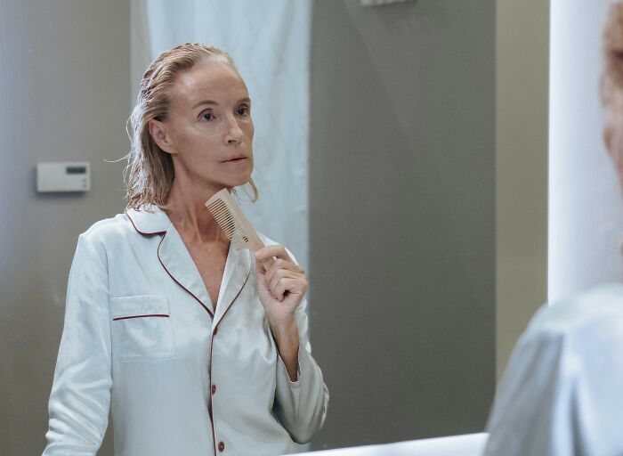 Middle-aged woman in white pajamas examining hair and body changes that indicate aging in a bathroom mirror.
