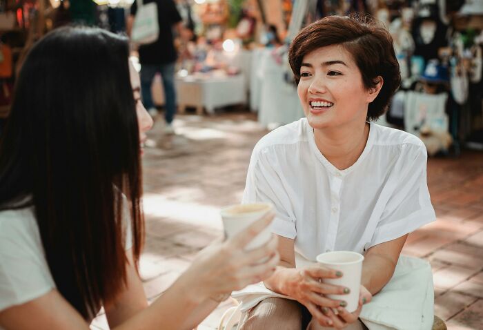 Two women laughing and chatting while holding cups outdoors, illustrating hilarious and weird things found secretly doing.