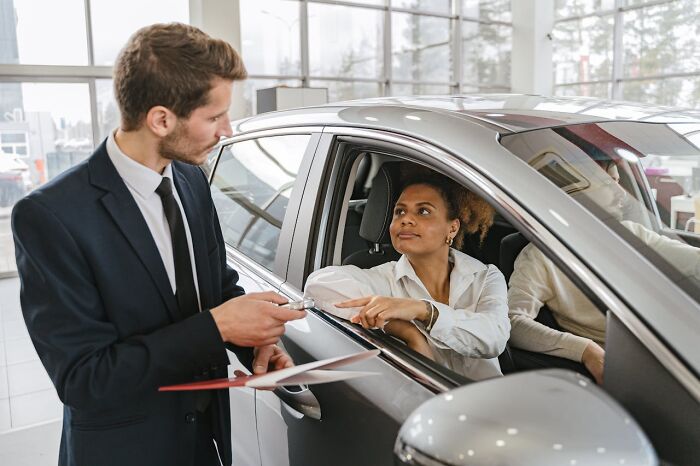 Car salesperson in suit talking to female customer in car, illustrating jobs that shouldn’t be earning as much as they do.