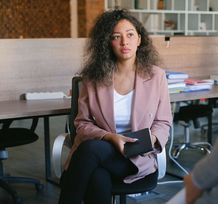 Young woman in a pink blazer sitting in office chair holding a notebook, representing real-life cheat codes for success.