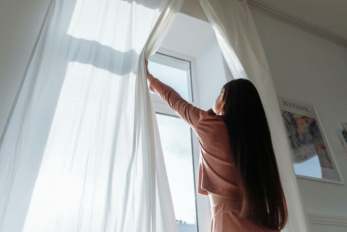 Woman enjoying low-key pleasures opening white curtains to let natural sunlight into a cozy, bright room.
