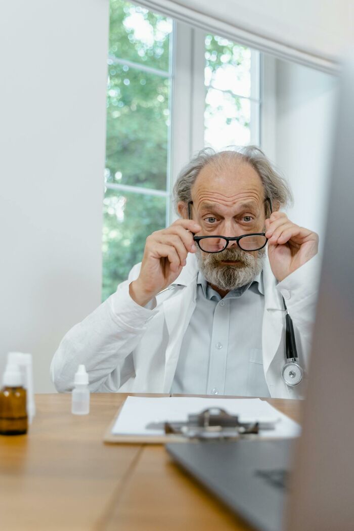 Elderly male doctor adjusting glasses in a bright office, representing weird and unhinged finds shared by medical professionals.