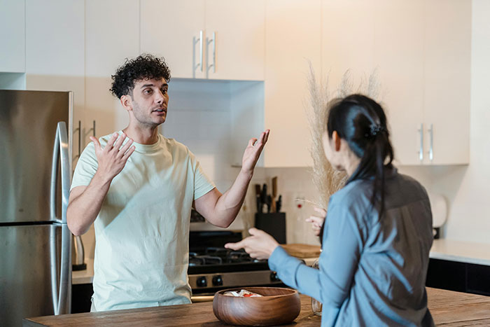 Man guarding box of olives at home, gesturing in frustration while woman cleans nearby in kitchen setting.
