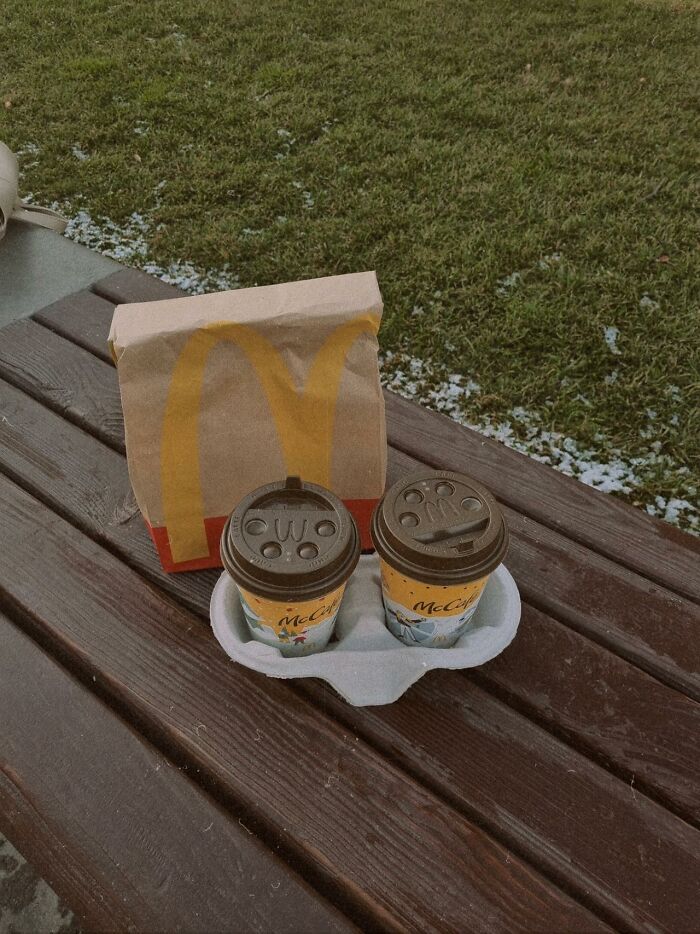 Two coffee cups in a holder and a fast food paper bag on a wooden bench, representing restaurant food items.