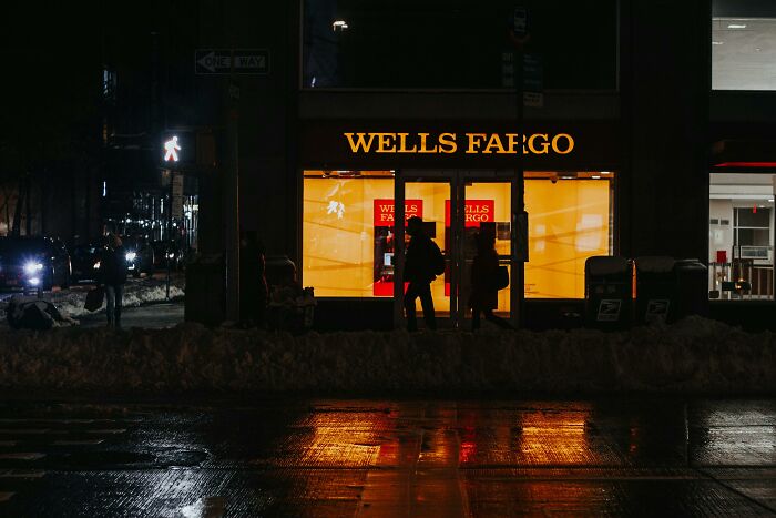 People share companies bad list as silhouettes stand outside a Wells Fargo branch at night on a wet city street.
