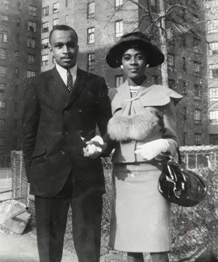 Vintage photo of a well-dressed couple standing together outdoors in front of apartment buildings, capturing history up close.