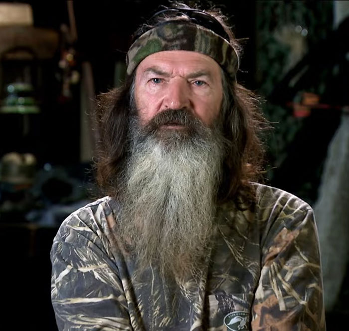 Phil Robertson from Duck Dynasty wearing camo shirt and headband with long beard in an indoor setting.