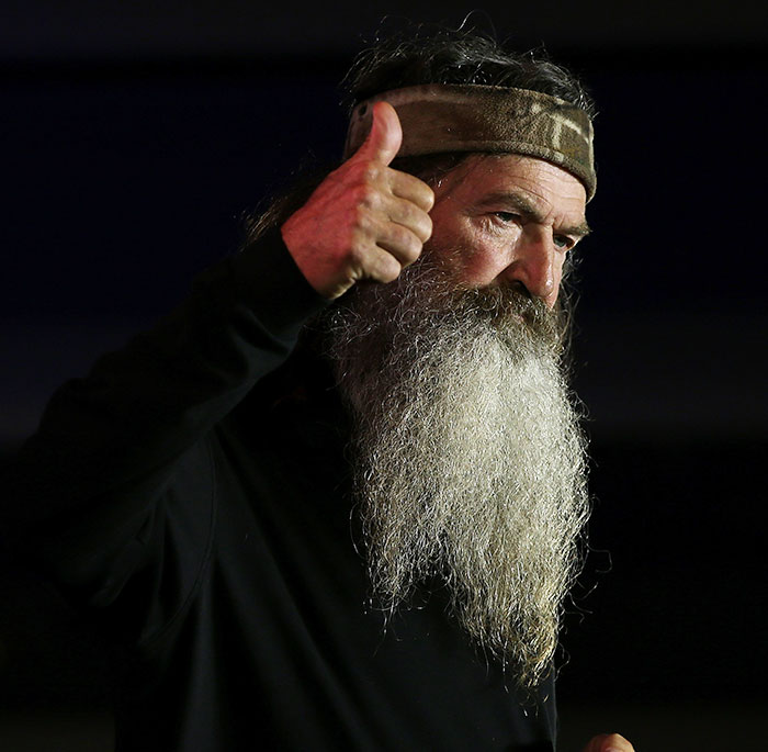 Phil Robertson from Duck Dynasty giving thumbs up, wearing a headband and black shirt against dark background.