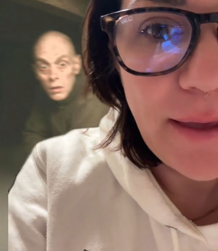 Woman in glasses and white hoodie shares story after spotting stranger phrogging and hiding under hotel bed.