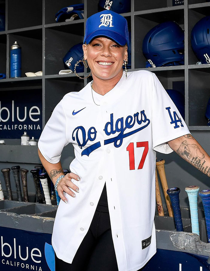 Smiling woman wearing a Dodgers jersey and cap posing in a baseball dugout with bats and helmets behind her.