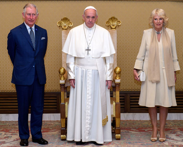 Pope in white robes seated between a man in a dark suit and a woman in a cream white outfit during a formal Vatican meeting.