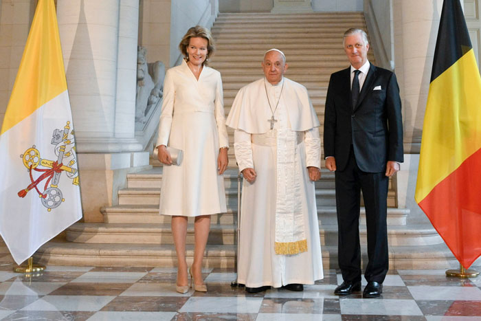 Woman in white outfit standing next to the Pope and a man between Vatican and Belgian flags in grand hall.