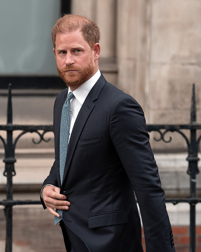 Prince Harry in a dark suit and patterned tie, looking serious and walking near a black wrought iron fence.