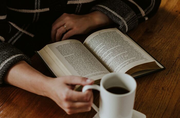 Person enjoying one of 36 low-key pleasures, reading a book while holding a cup of coffee at a wooden table.