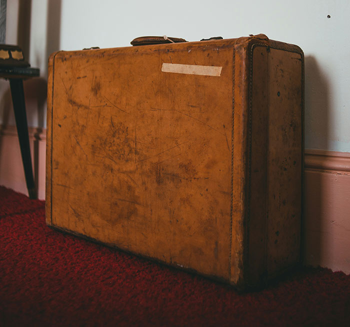 Worn brown leather suitcase on red carpet near wall, symbolizing emotional baggage in siblings of narcissists and sociopaths