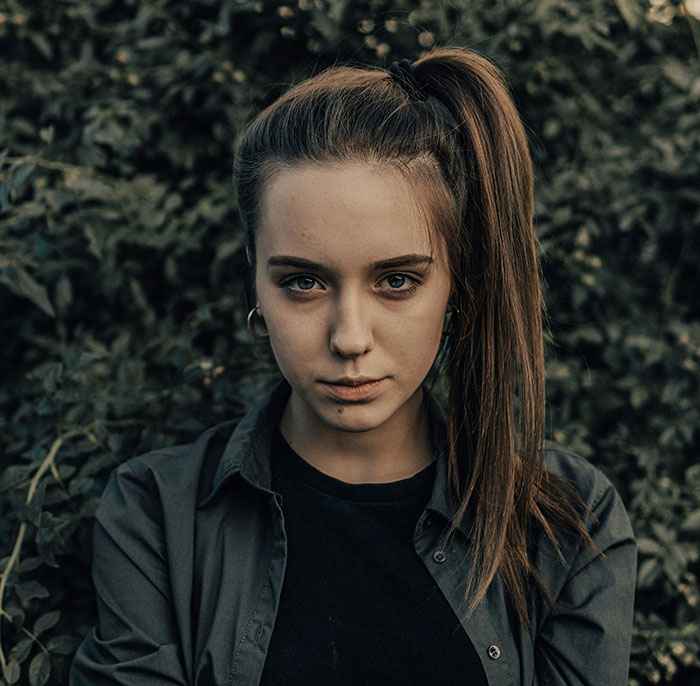 Young woman with serious expression standing outdoors surrounded by foliage, reflecting on siblings of narcissists and sociopaths.