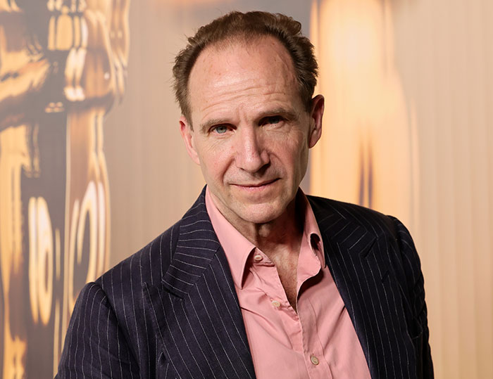 Ralph Fiennes in a dark pinstripe suit and pink shirt posing indoors, linked to reunion with Francesca Annis after affair.
