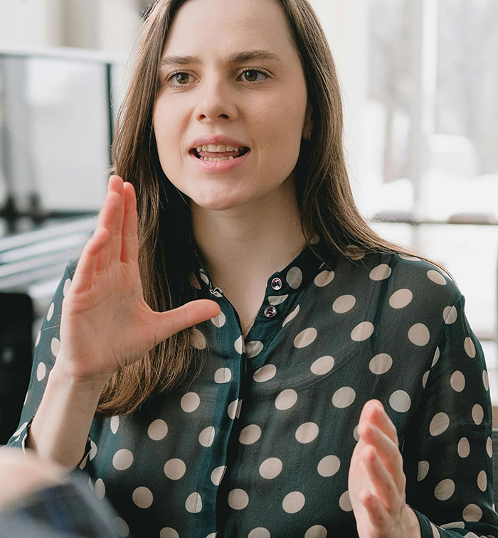 Young woman with brown hair wearing a polka dot shirt, gesturing while explaining a funny moment about wind made by trees.