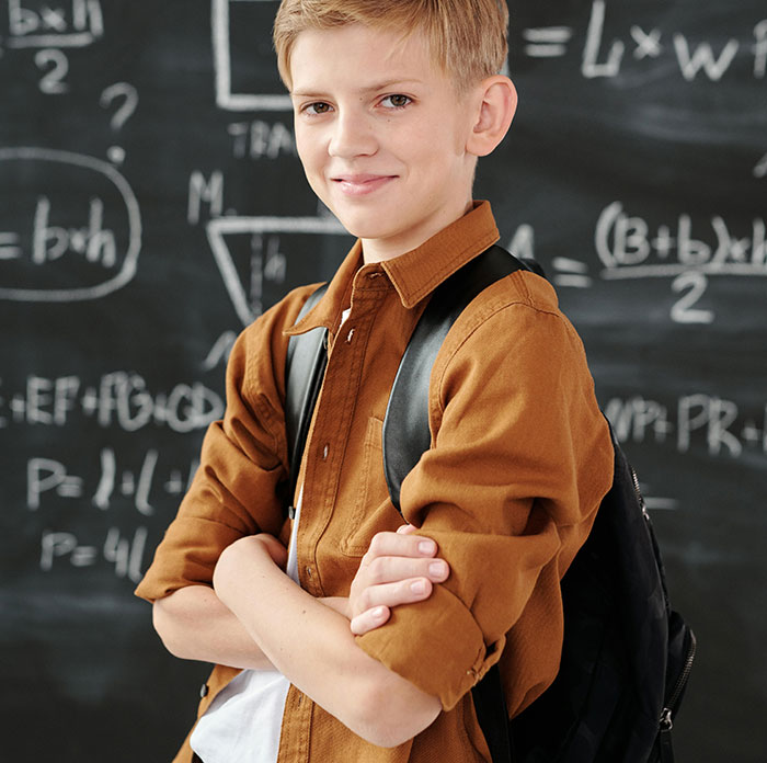 Confident schoolboy with backpack standing in front of a chalkboard filled with math formulas, illustrating huge risks concept.