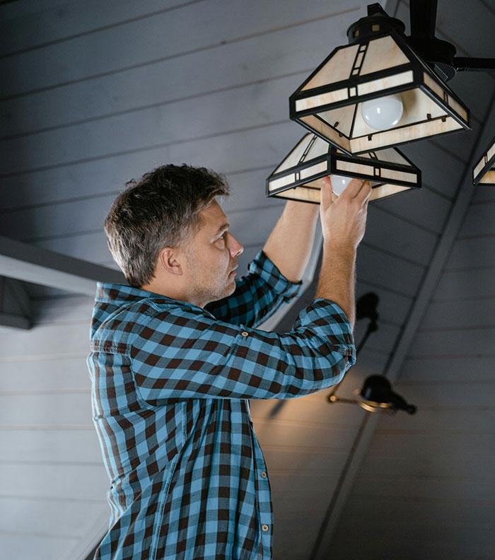 Man in a blue plaid shirt taking a huge risk while changing a ceiling light fixture in a high attic room.