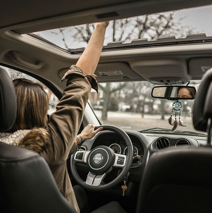Woman driving Jeep with sunroof open, reaching upward, with trees visible outside and dreamcatcher hanging from rearview mirror.