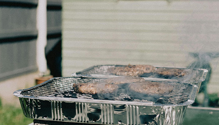 Portable grill with burgers cooking outdoors on a sunny day, smoke rising as wind is made by trees.