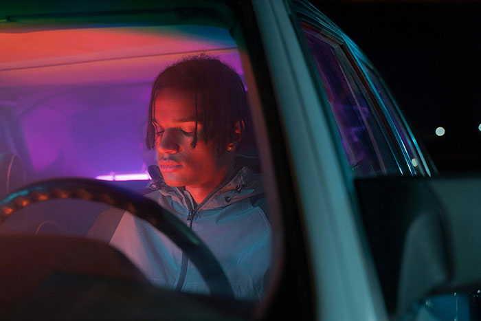 A young man inside a car at night illuminated by colorful lights, illustrating people taking huge risks for no logical reason.