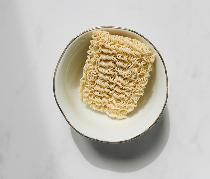 Square block of uncooked instant ramen noodles in a white bowl on a light gray surface, emphasizing simple food items.