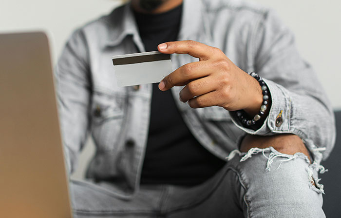 Person in ripped jeans holding a credit card near a laptop, illustrating huge risks taken with no logical reason.