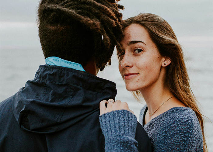 Young woman looking at camera while holding partner's shoulder by the sea, illustrating stories of parents disowning their kids.