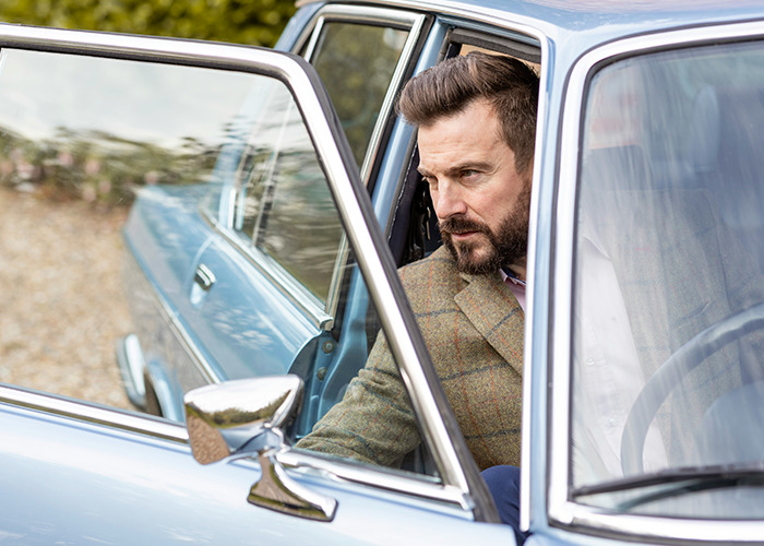 Man with beard sitting in vintage car, looking serious and contemplative, reflecting on parents disowning their kids stories.